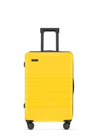 Valise rigide jaune avec un design élégant à stries, poignée noire rétractable et quatre roues noires pour une mobilité facile.