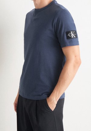 T-shirt en coton bleu marine avec un col rond côtelé, des manches courtes et un écusson noir sur la manche gauche. Texture lisse, coupe régulière.