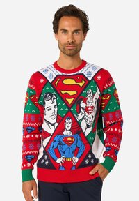 Muž v farebnom vianočnom svetri so vzorom Supermana, na ktorom sú viaceré komiksové ilustrácie Supermana a vzory snehových vločiek.