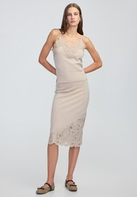 Brede twee-delige set in lichtbeige, met complexe kantafwerking op de top en de zoom. Slank figuur met spaghettibandjes en een aangesloten rok.