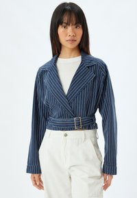 Marinrandig kort jacka med stora slag, bälte i midjan och långa ärmar. Tillverkad av ett texturerat tyg. Matchas med ljusa byxor.