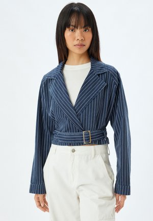 Marineblaue gestreifte Kurzjacke mit großen Revers, tailliertem Gürtel und langen Ärmeln. Hergestellt aus einem strukturierten Stoff. Kombiniert mit hellen Hosen.