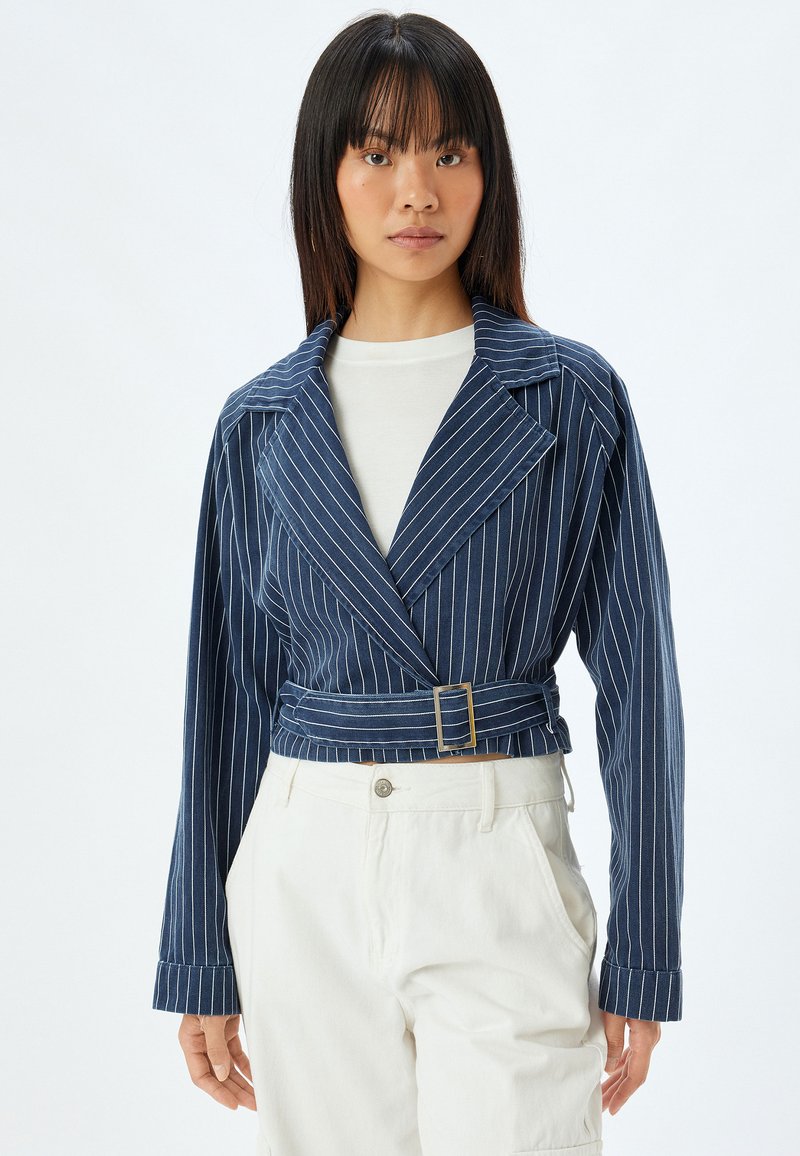 Marinrandig kort jacka med stora slag, bälte i midjan och långa ärmar. Tillverkad av ett texturerat tyg. Matchas med ljusa byxor.