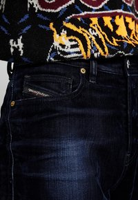 Jeans in denim blu scuro con una leggera lucentezza, caratterizzati da una tasca con un'etichetta rosso e bianco di Diesel Industries e rivetti dorati come dettagli.