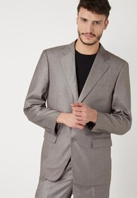 Chaqueta de traje gris claro con solapas de muesca, cierre de un solo botón, dos bolsillos frontales y tela texturizada. Llevada sobre una camisa negra.