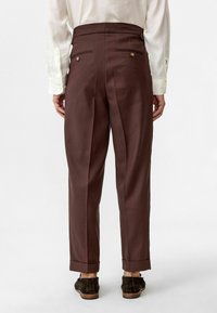 Bruine, op maat gemaakte broek met een middelhoge taille, plooien en twee achterzakken, voorzien van goudkleurige knopen en opgerolde mouwen. Draagt met een licht shirt.
