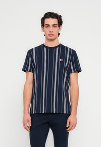 Μπλουζάκι από βαμβάκι σε ναυτικό μπλε με κάθετες λευκές ρίγες, κοντά μανίκια και μικρό λογότυπο στην αριστερή πλευρά στο στήθος. Σχεδιασμός με στάνταρ στρογγυλή λαιμόκοψη.