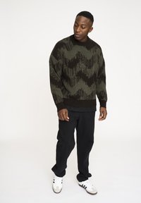 Dicker Strickpullover in Olivgrün mit schwarz-weißer Zickzack-Musterung, runder Ausschnitt, gerippte Bündchen, über einem schwarzen Shirt getragen, kombiniert mit schwarzen Hosen und weißen Sneakers.