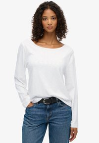 Witte top met lange mouwen en een ruime halslijn, gecombineerd met blauwe jeans en een zwarte riem. Glad textuur en een ontspannende pasvorm.