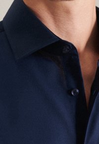 Mörkblå button-up skjorta med en strukturerad krage, slät tygtextur och en enda knappdetalj vid halsen.