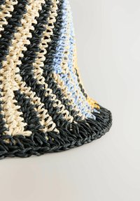Panier tissé mettant en avant des fibres noires, beiges, bleu clair et jaunes tressées en motif en zigzag, avec un bord à franges et une finition texturée.