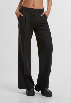 Persoon draagt zwarte wijde broek met hoge taille en zijzakken en zwarte stevige plateauzolen, staat met handen in de zakken.