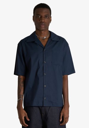 Giovane uomo con camicia blu navy a maniche corte con bottoni e tasca frontale e pantaloni scuri su sfondo semplice.