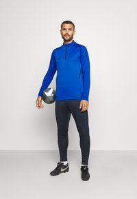 Blauw sportshirt met lange mouwen en ritskraag, zwarte accenten en logo, gecombineerd met een donkere broek met een blauwe streep en knopen. Houdt een voetbal vast.