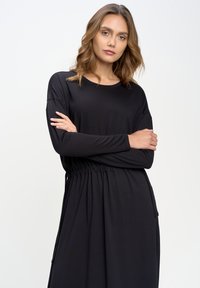 Juoda ilga rankovė su apvalia iškirpte ir elastinga juosmens linija. Pagaminta iš lengvos, lygios medžiagos; turi šonines kišenes.