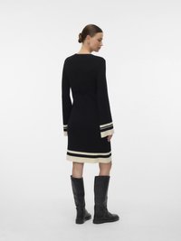 Nő háttal áll, fekete térd alá érő ruhát visel, fehér csíkokkal az ujjain és az alján, fekete térdig érő csizmával párosítva.