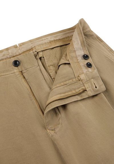 Beige broek met een open rits, twee zwarte knopen op de tailleband en een omgevouwen taillebandflap op een witte achtergrond.