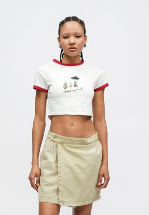 Λευκό crop top με κόκκινο τελείωμα, διακοσμημένο με πολύχρωμο γραφικό σχέδιο. Μπεζ φούστα τύπου περικάλυψης με κούμπωμα κουμπιού και υφή υφάσματος.