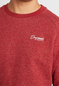 Sudadera roja con un patrón texturizado, con cuello redondo y logotipo bordado en blanco "Originals" en el lado izquierdo.