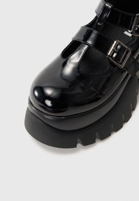 Sapatos plataforma de лаquer preto com bico arredondado, tiras no tornozelo e sola de borracha grossa com uma borda serrilhada.