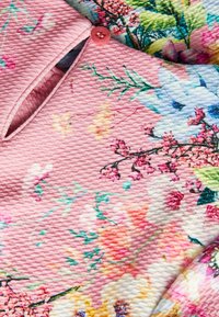 Vaaleanpunainen kukkainen paita, jossa on tekstuuri ja kaulus, jota korostaa pieni vaaleanpunainen nappi. Kirkkaat värit kukkakuoseissa ovat myös näkyvissä.