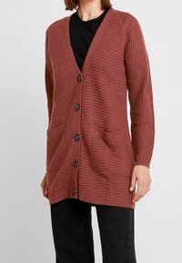Langer, rostfarbener Strickcardigan mit V-Ausschnitt, sechs schwarzen Knöpfen, zwei Fronttaschen und gerippten Bündchen. Strukturierter Stoff.