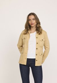 Veste en coton beige avec un devant à boutons, deux poches poitrine et des boutons contrastants. Portée au-dessus d'une chemise blanche et associée à un jean noir.