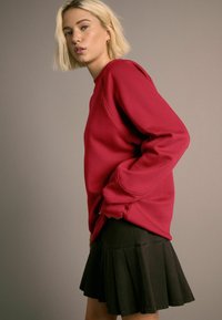 Punainen huppari, jossa on pitkät hihat, pyöreä kaula-aukko ja rento leikkaus, yhdistettynä mustiin farkkuhameeseen, jossa on laskostettu helma.