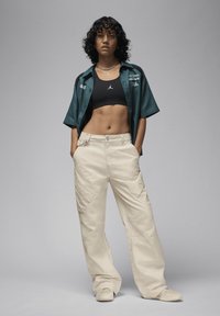 En model bærer en sort crop top og en teal kortærmet skjorte med broderede detaljer, parret med beige cargo bukser med sidelommer.