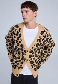 Cardigan in stampa leopardata in morbido tessuto beige, con macchie nere e marroni, scollo a V e tasche frontali con chiusure a bottone.