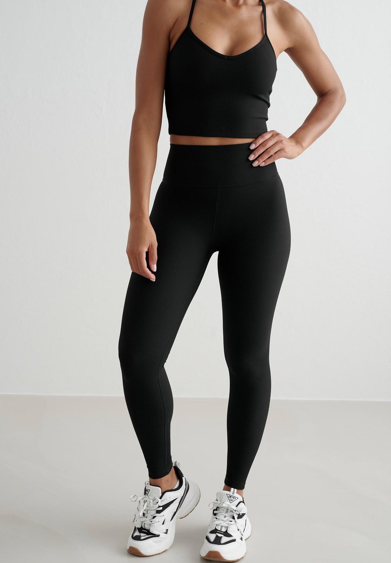 Schwarzes geripptes Cropped Top kombiniert mit hochtaillierten schwarzen Leggings aus glattem Material. Gestylt mit weißen und schwarzen Sportschuhen.