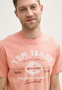 Camiseta de color coral claro con cuello redondo y detalles de logotipo impresos en blanco. Tejido de algodón suave con un corte clásico y sin patrones visibles.