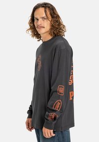 Černé tričko s dlouhým rukávem a oranžovými grafickými potisky na rukávech a přední části. Měkký bavlněný materiál s uvolněným střih a kulatým výstřihem.