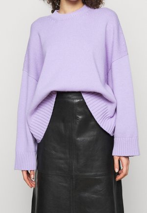 Kvinde iført en lavendelstrikket sweater med oversize ærmer, stoppet ned i et sort lædernederdel med høj talje.