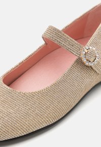 Zapato tipo Mary Jane de tela dorada con punta redonda, acabado texturizado, pequeña correa y un acento de joya circular en la correa, forrado en rosa.