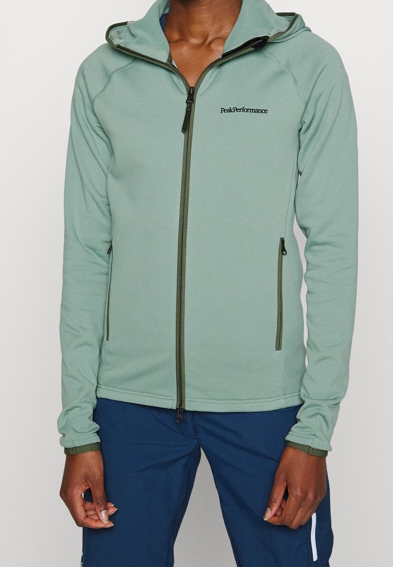 Veste polaire vert clair avec capuche. Elle comprend une fermeture éclair à l'avant, des poches latérales et un logo sur le côté gauche de la poitrine. Texture lisse et coupe ajustée.