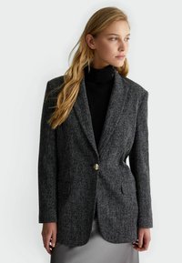Blazer grigio texturizzato con maniche lunghe, chiusura a un bottone dorato e due tasche frontali. Indossato sopra un dolcevita nero.