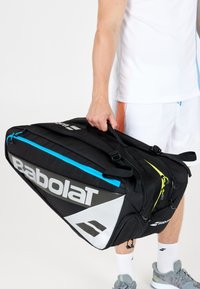 Persona in abbigliamento sportivo bianco che tiene in mano una grande borsa da tennis Babolat nera con dettagli della cerniera blu e gialla.