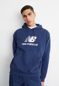 Tengerészkék kapucnis pulóver, puha anyagból készült, elől zsebbel, húzózsinórral ellátott kapucnival és nagy fehér New Balance logóval a mellkason.