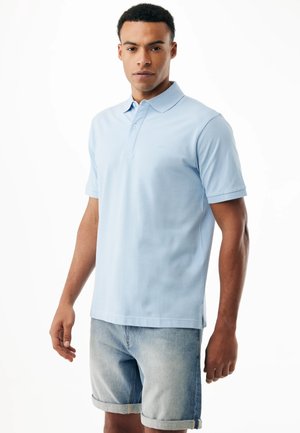 Mann steht, trägt ein hellblaues Polohemd und hochgekrempelte Jeansshorts vor einem schlichten weißen Hintergrund.