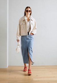 Beige cropped jacket with two pockets, white shirt, light blue denim skirt with front slit, and red sneakers, holding a white handbag.