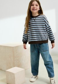 Leende tjej klädd i en blå och brun randig tröja, löst sittande denimjeans och vita sneakers med svarta mönster, stående vid beige kuber.