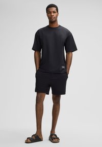 Junger Mann steht mit den Händen in den Taschen, trägt ein übergroßes schwarzes T-Shirt, schwarze Shorts und schwarze geschnallte Sandalen vor hellen Hintergrund.