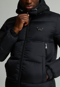 Schwarze Pufferjacke mit Reißverschluss, gefaltetem Kapuzenkragen und Logodetail. Verfügt über gestepptes Material und seitliche Reißverschlusstaschen. Glatte, glänzende Textur.