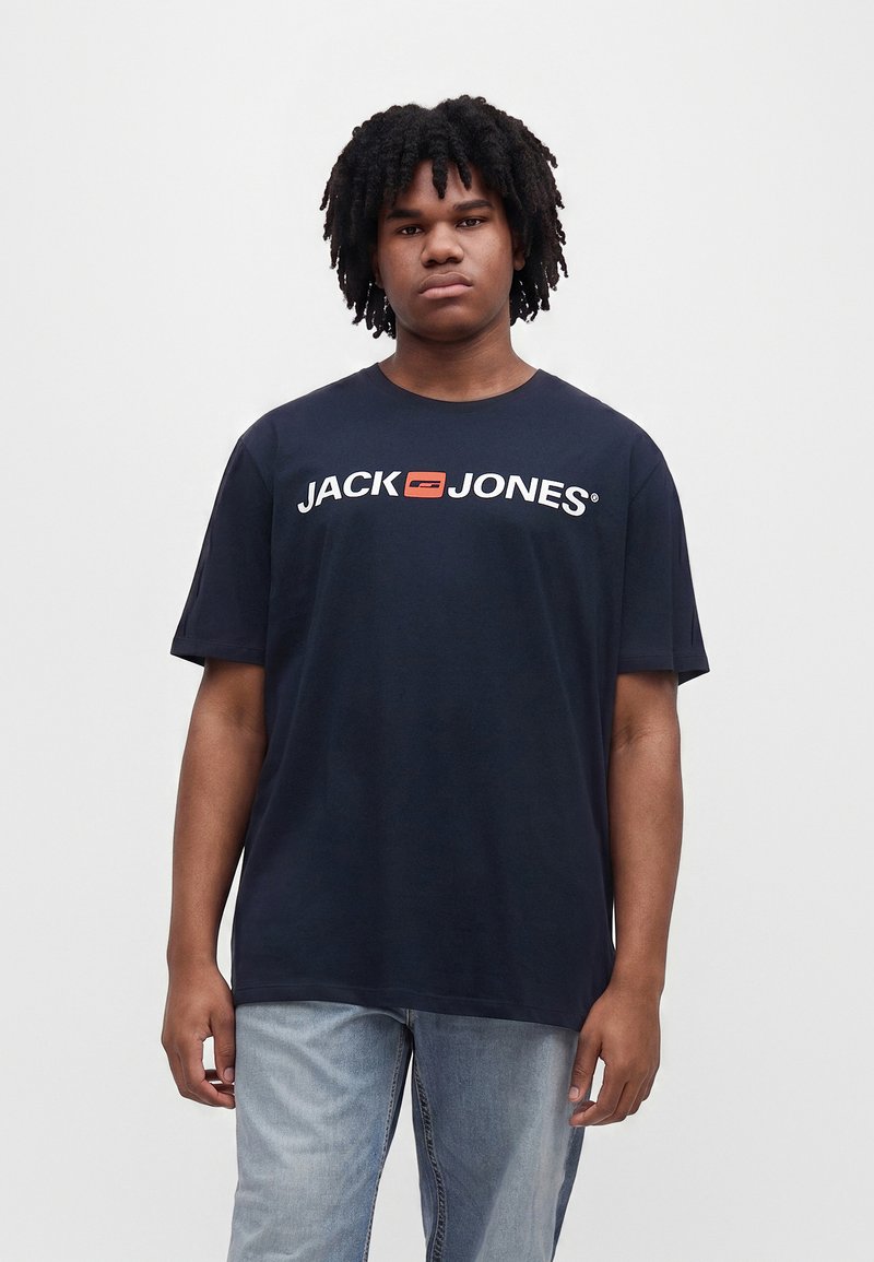 Młody mężczyzna z krótkimi kręconymi włosami, ubrany w granatowy t-shirt Jack Jones i jasnoniebieskie jeansy, stoi na jednolitym tle.