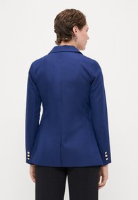 Laivansininen räätälöity bleiseri, jossa on teksturoitu kangas, leikattu kaulus ja kultasävyiset nappikoristeet hihoissa. Istuu vartalonmyötäisesti ja on kellotettu muotoilultaan.