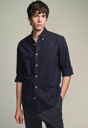 Navy blue button-up shirt made of cotton, featuring long sleeves rolled up, a front pocket, and white buttons. Smooth texture and classic design.
