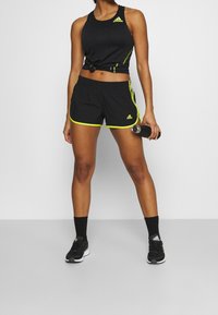 Camiseta sin mangas deportiva negra y pantalones cortos con detalles en neón amarillo. Calcetines y zapatos negros completan el look deportivo, sosteniendo una botella de agua.