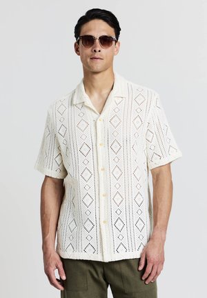 Chemise blanche à manches courtes avec un motif en losanges découpés et des boutons, portée avec des lunettes de soleil marron. Elle possède une coupe décontractée et une texture douce.