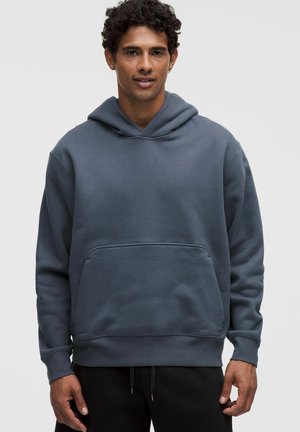 Mann mit lockigem Haar, der einen dunkelgrauen Kapuzenpullover und schwarze Hose trägt, steht vor einem einfarbigen hellen Hintergrund.